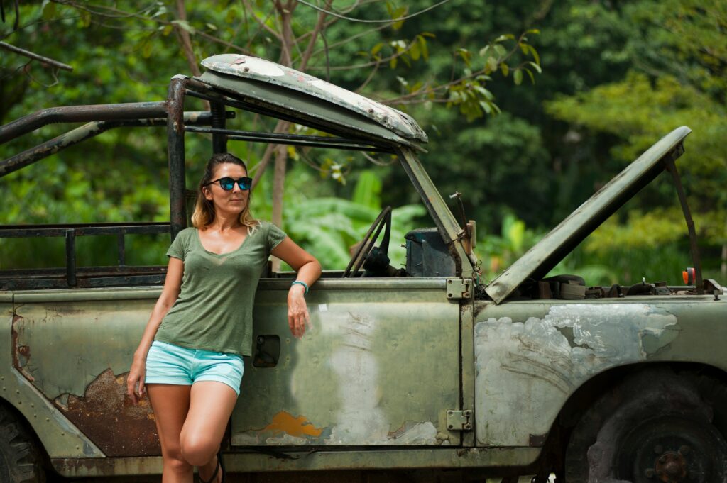 A woman leans against a rustic vintage car in a lush outdoor setting in Thailand.